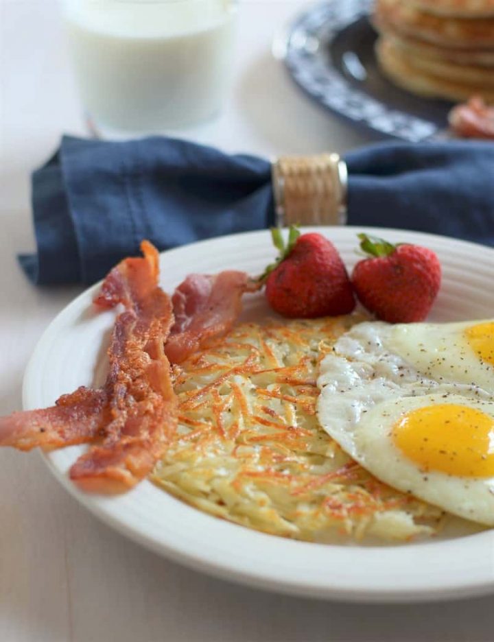 White plate with crispy golden shredded hashbrowns, 2 eggs sunny side up, 2 slices bacon, 2 strawberries with blue napkin and glass of milk