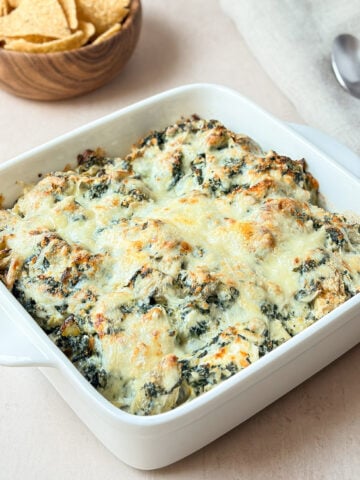 Creamy spinach artichoke dip baked in a white square dish with a golden cheesy top, served with tortilla chips.