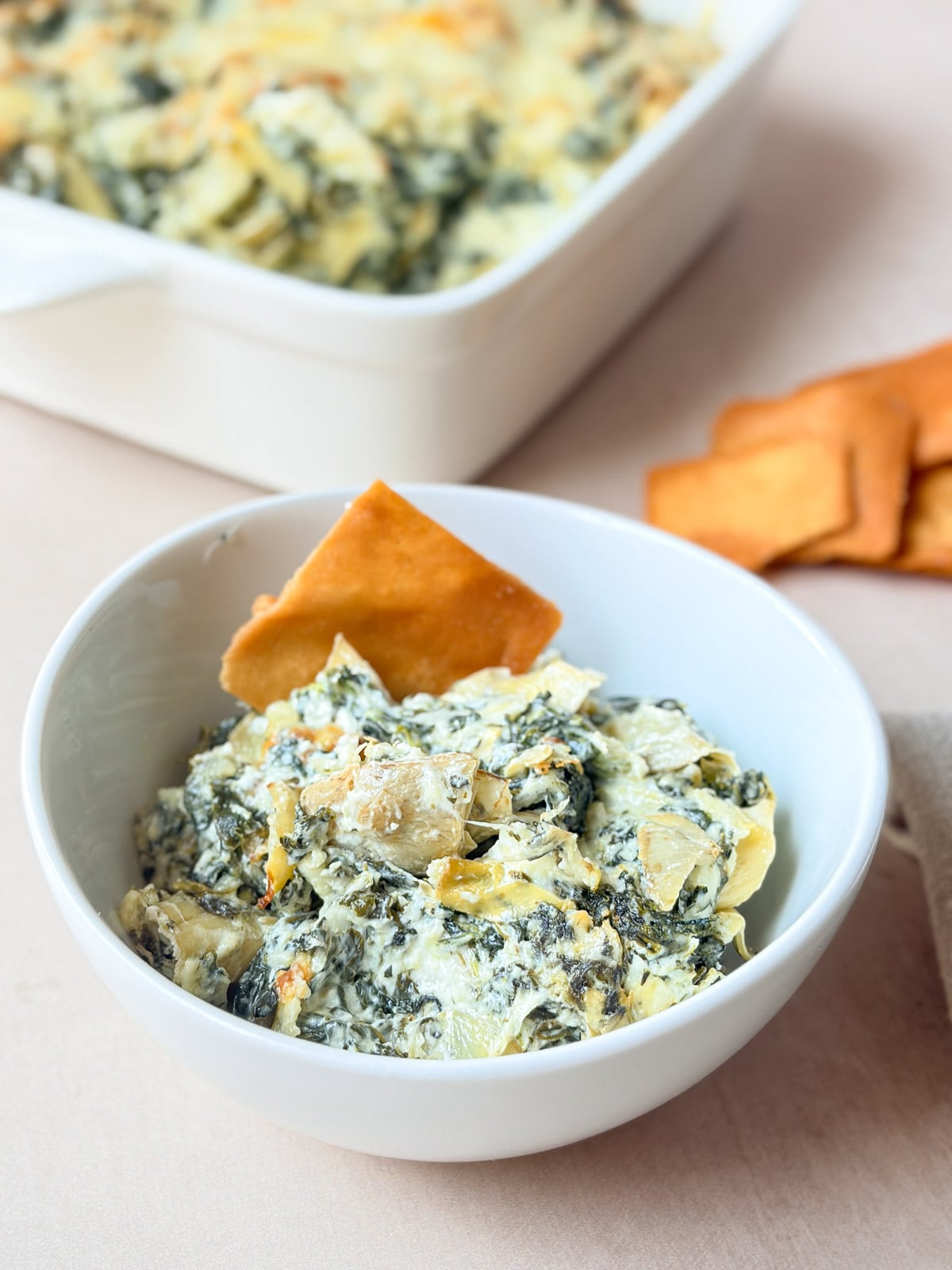 Spinach artichoke dip served in a white bowl with a tortilla chip and the baked dip in the background.