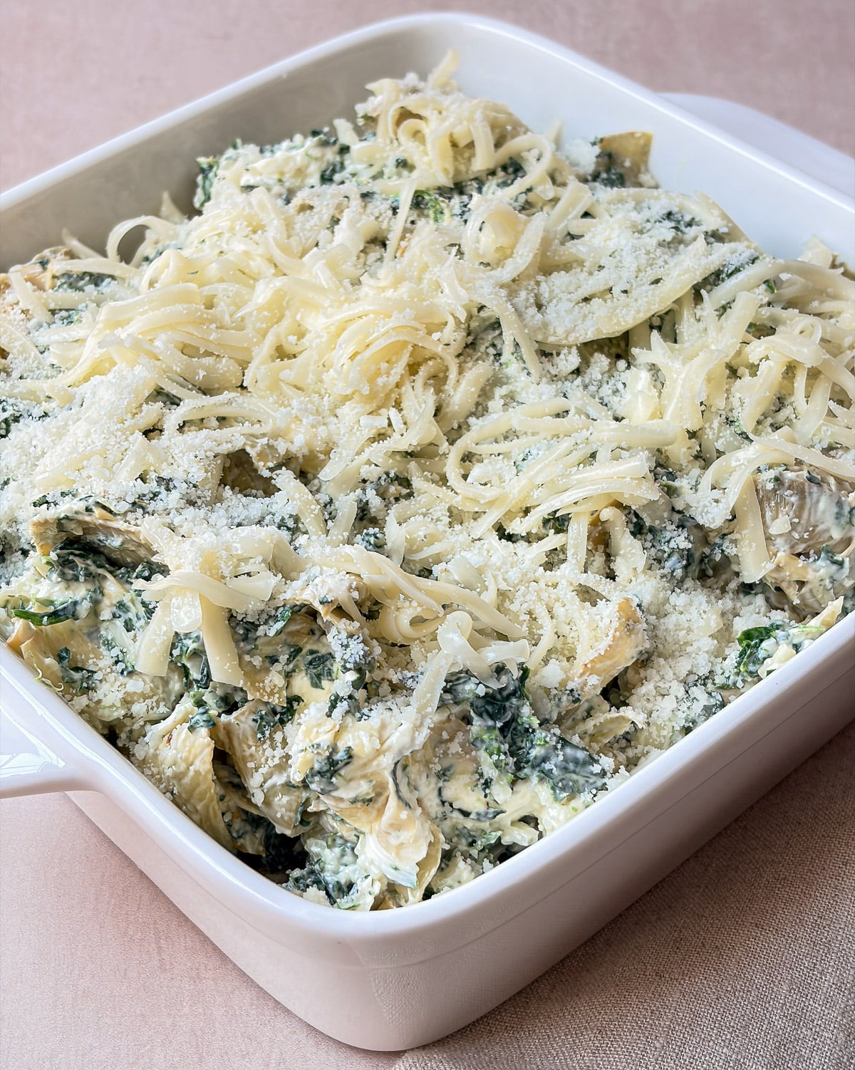 Unbaked spinach artichoke dip in a white square baking dish topped with shredded mozzarella and Pecorino Romano.