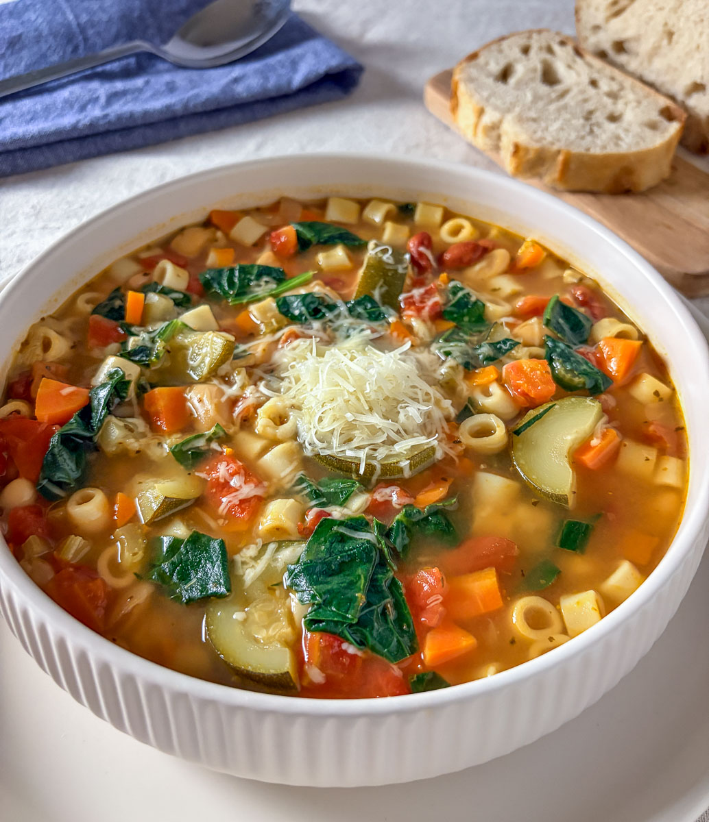 a bowl of minestrone soup with Parmesan cheese and bread in the background