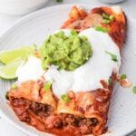 two ground beef enchiladas on a plate with sour cream, cheese and guacamole on top.