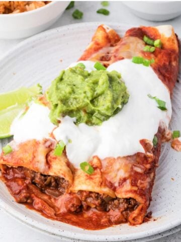 two ground beef enchiladas on a plate with sour cream, cheese and guacamole on top.