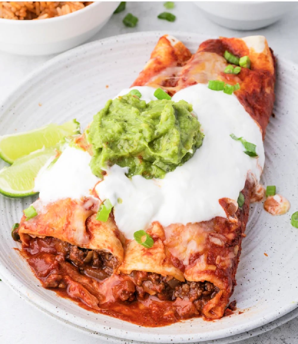 Two ground beef enchiladas on a plate with cheese, sauce, sour cream, and guacamole on top.