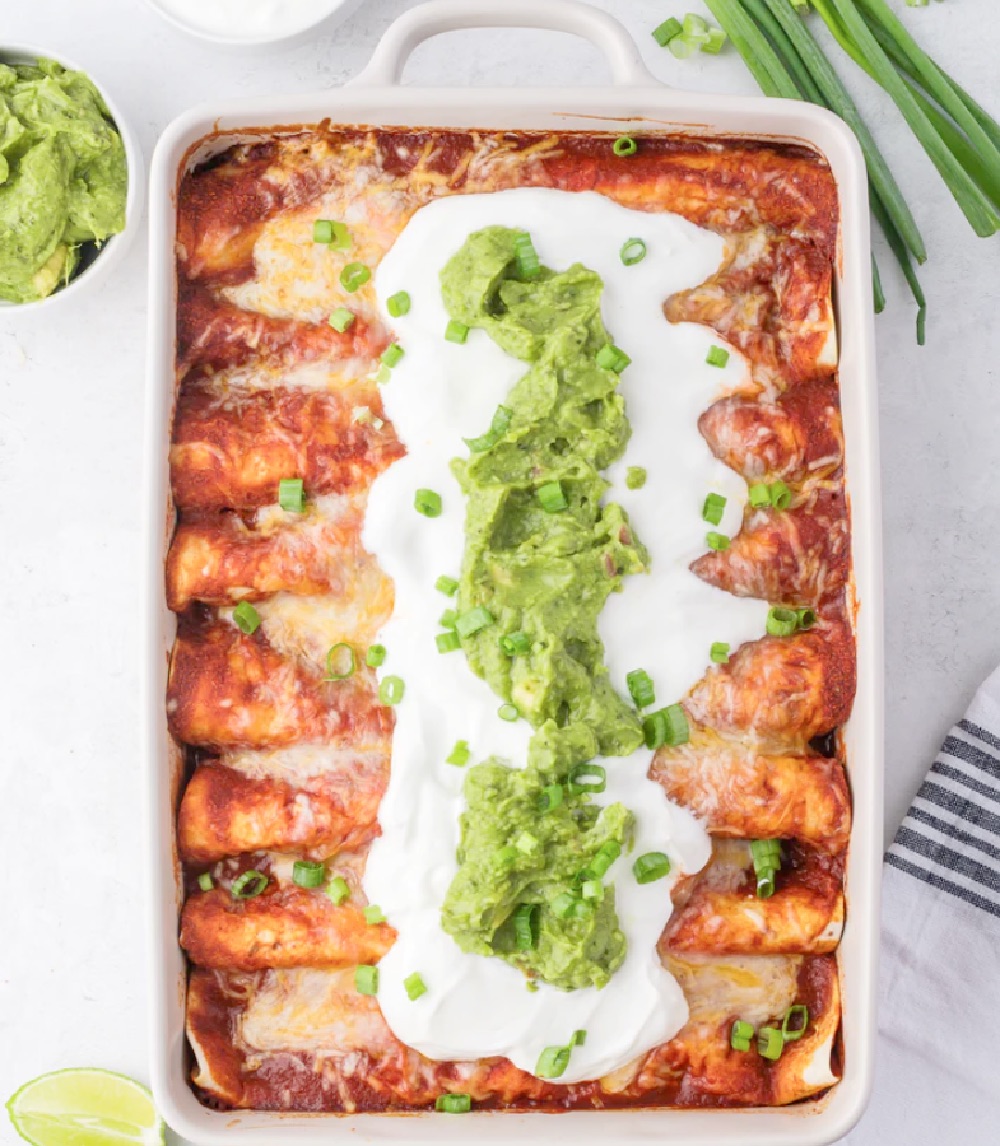 Ground beef enchiladas in a ceramic casserole dish with cheese, sour cream, chives and guacamole on top.