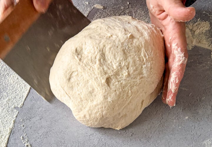 Sourdough being shaped on countertop with scraper and hand.